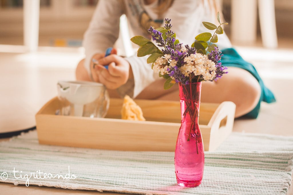 en segundo plano una niña aprendiendo con bandeja sensorial y jarrón traslucido rosa con flores en primer plano
